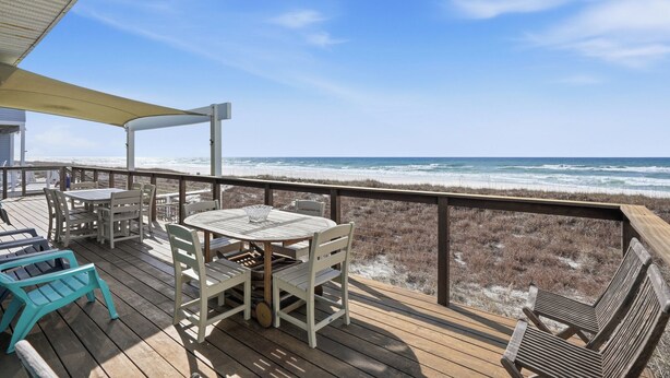A wooden deck with a table and chairs, overlooking a beach with waves.