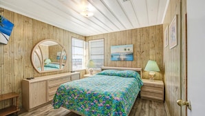 A bedroom with a wooden paneled wall, a bed with a floral bedspread, a round mirror, two bedside tables with lamps, and a painting on the wall.