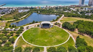 An aerial view of a park with a central stage, surrounded by a circular lawn, a lake, and a beach area.