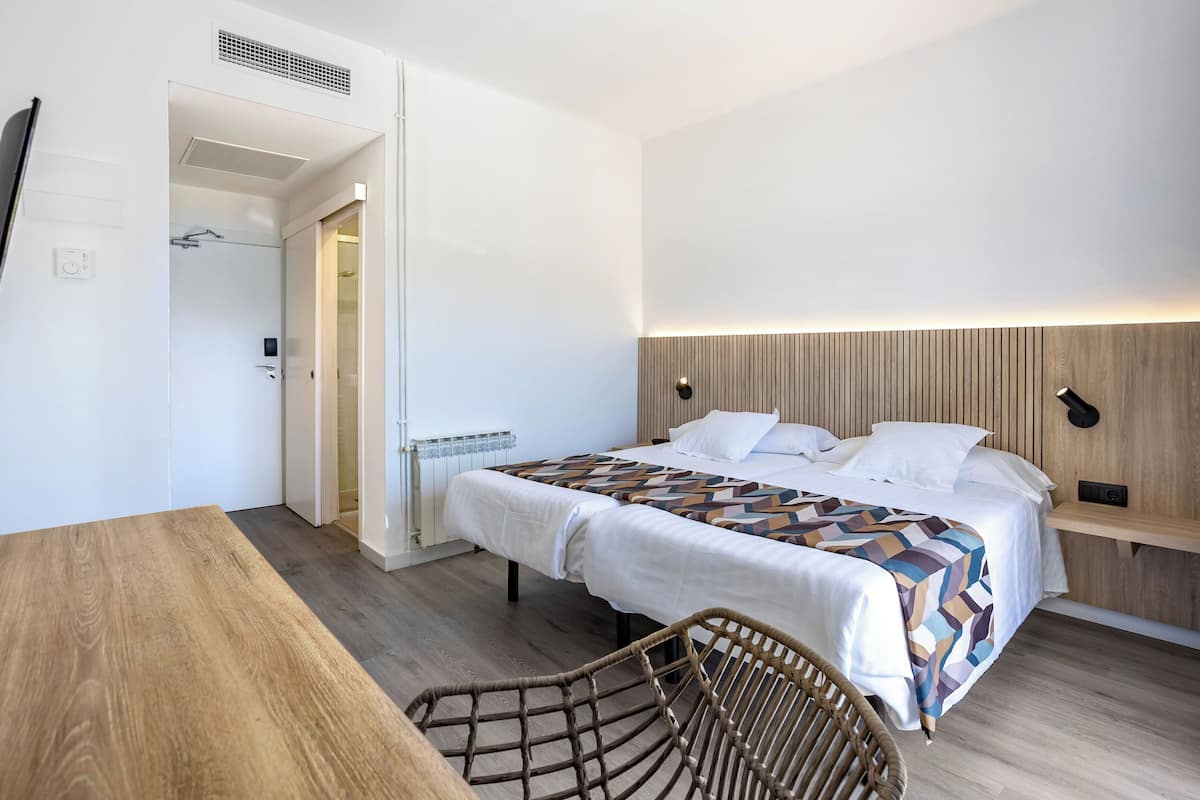 A modern hotel room with a wooden floor, a bed with a patterned bedspread, a bedside table with a lamp, and a wicker chair.