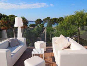 A terrace with white outdoor furniture, a glass railing, and a view of the sea and trees. - Hotel Morlans Garden (Calvia)