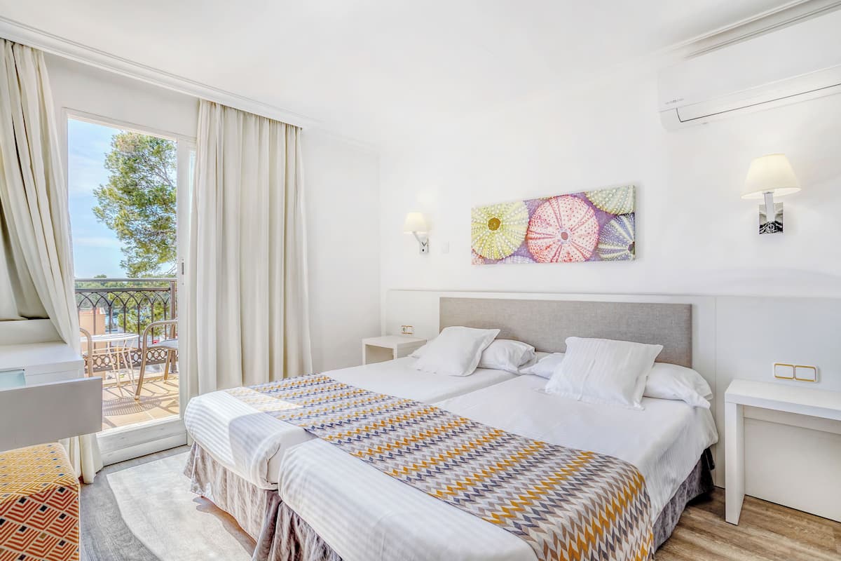 A hotel room with a bed, a desk, a chair, a window with curtains, a wall-mounted lamp, and a decorative wall piece.