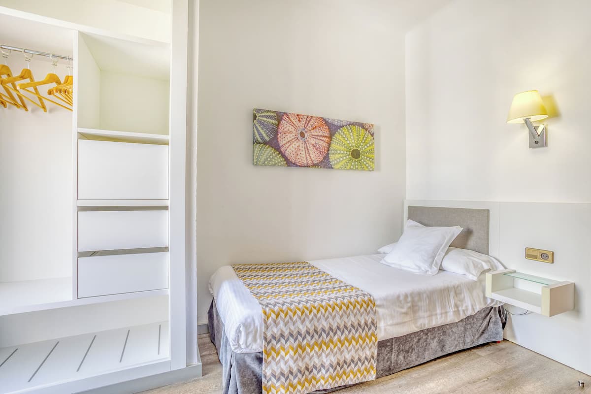 A bedroom with a bed, a bedside table, a wall-mounted lamp, a shelf, and a closet with hanging clothes.
