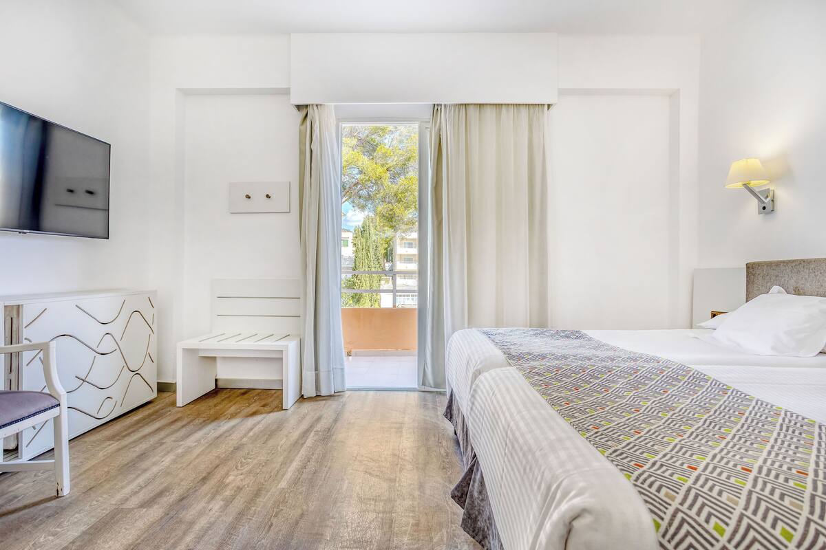 A hotel room with a bed, a desk with a chair, a TV, and a balcony with curtains.