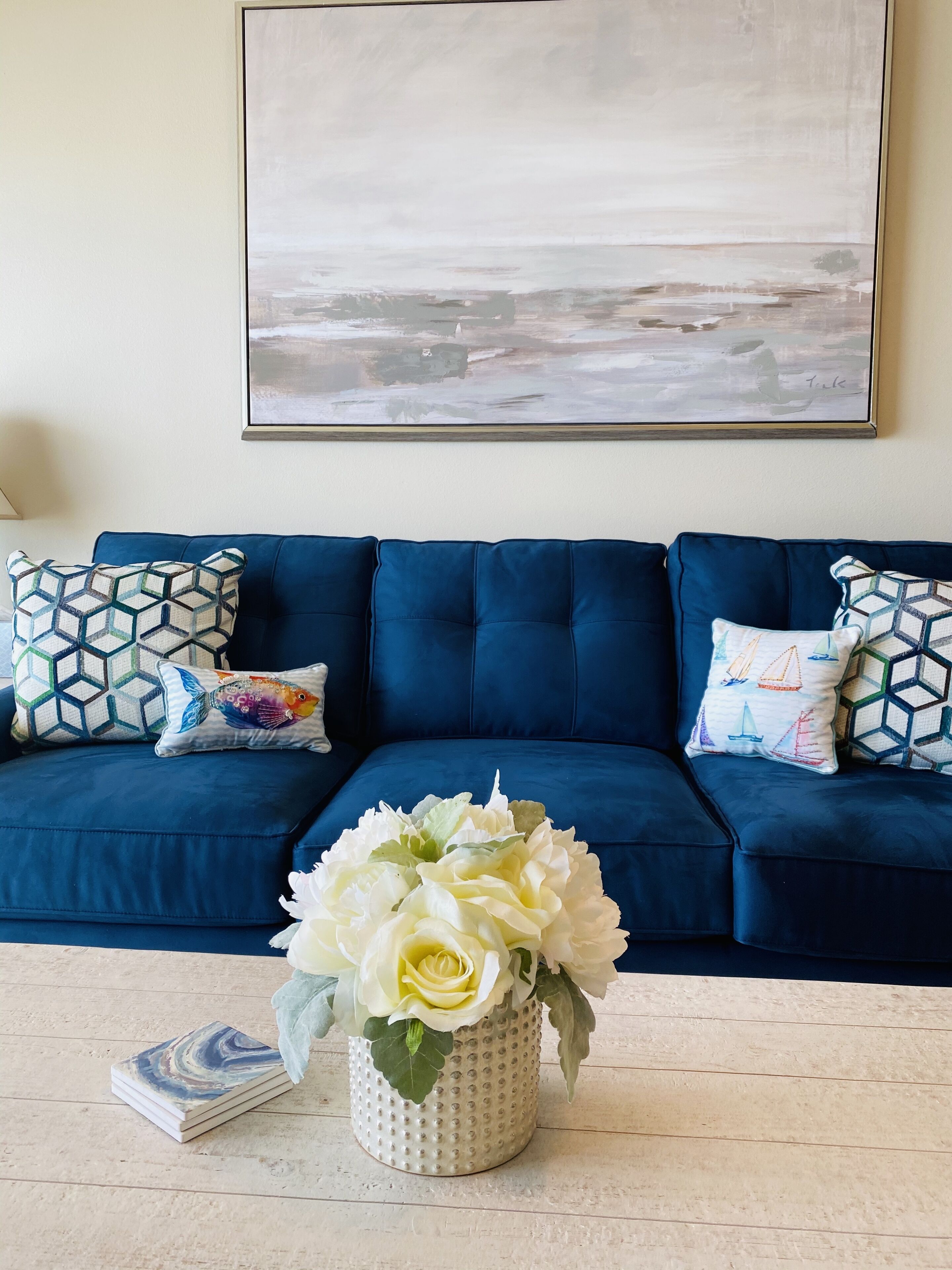 A blue sofa with patterned cushions, a white vase with flowers, and a framed abstract painting on the wall.