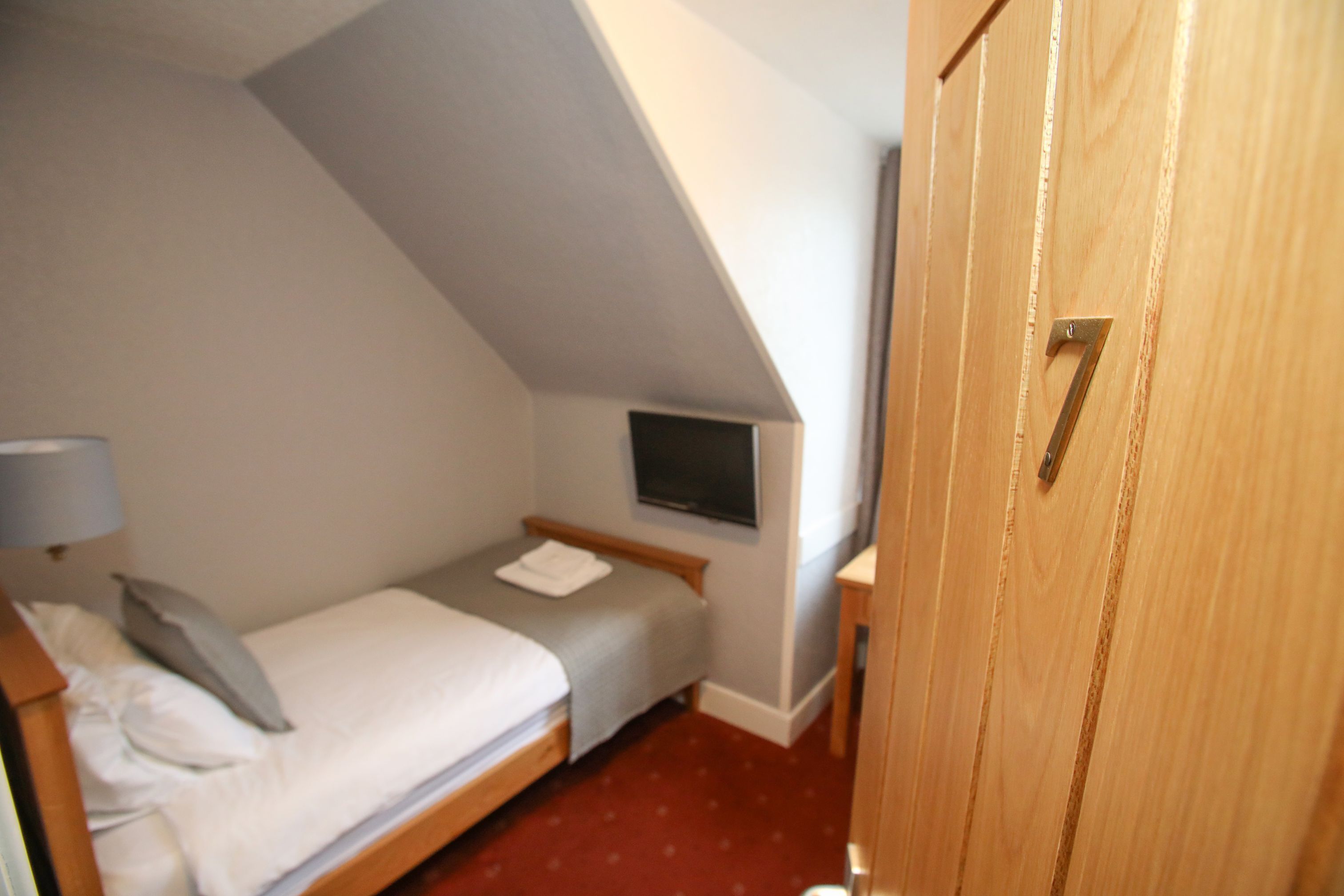 A small, single-bed room with a sloped ceiling, a television mounted on the wall, and a wooden door with a number.