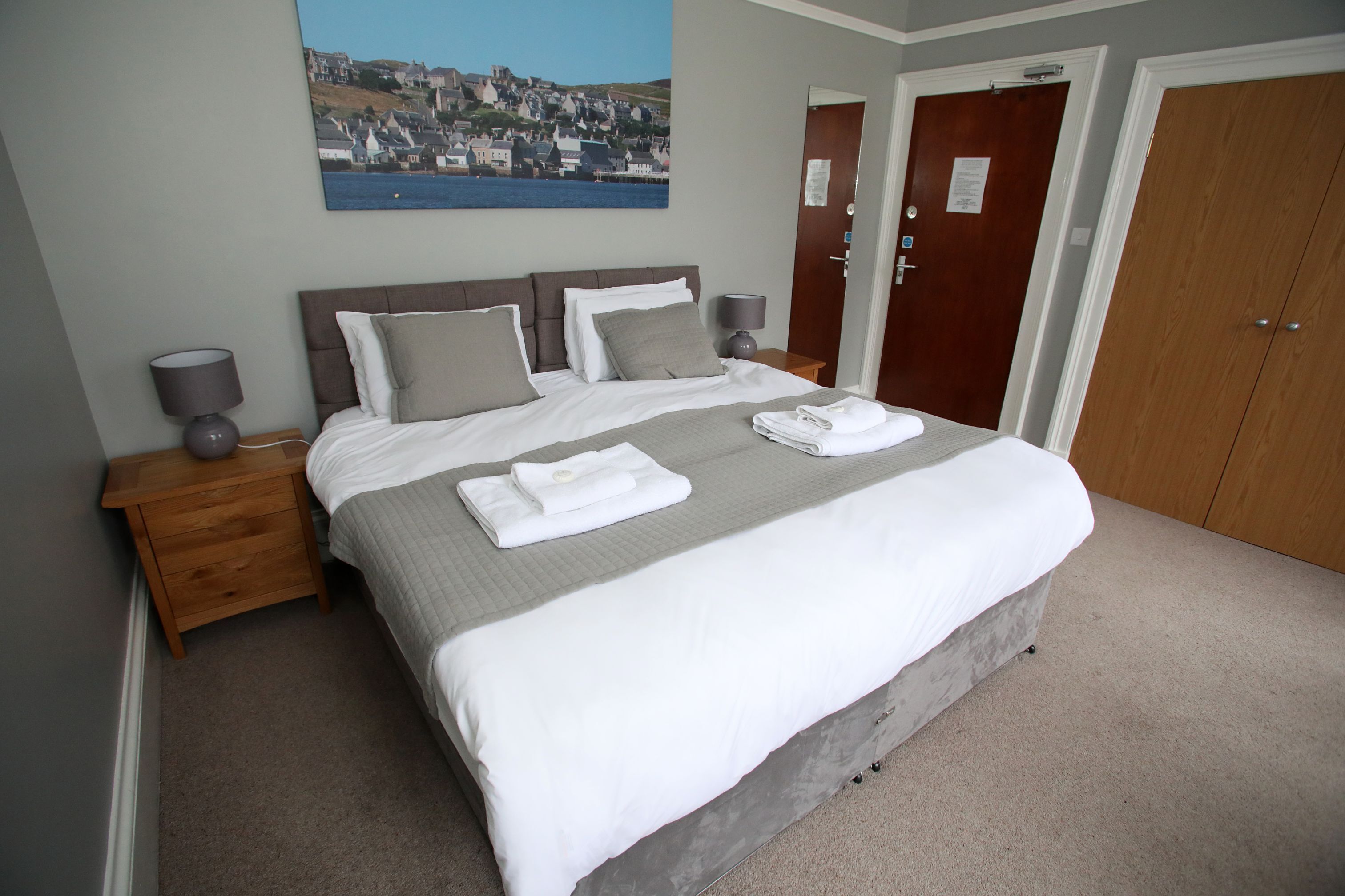 A neatly made bed with white linens and gray pillows, a wooden nightstand with a lamp, and a painting of a coastal town on the wall.