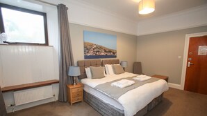 A bedroom with a large bed, a window with curtains, a radiator, a bedside table with a lamp, and a painting on the wall. - The Royal Hotel Stromness (Stromness)