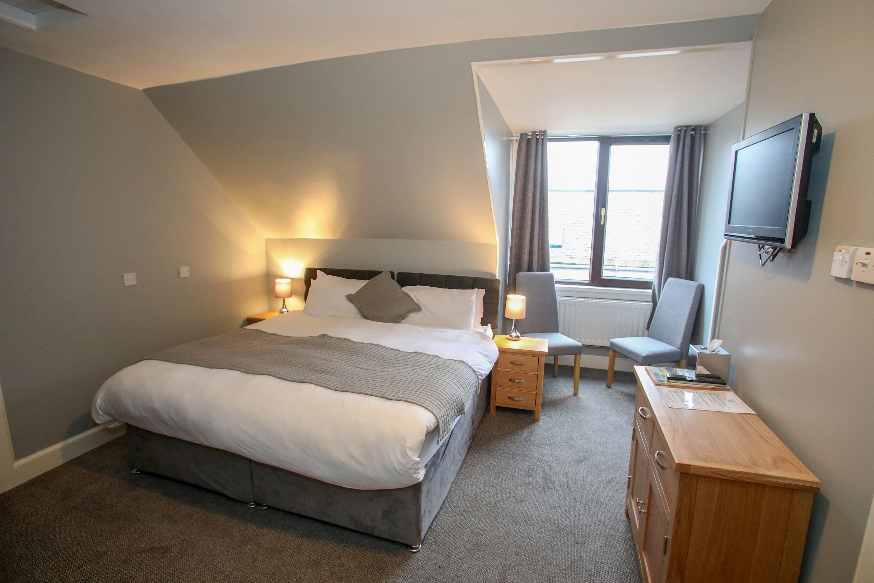A hotel room with a large bed, bedside lamps, a desk with a chair, a TV, and a window with curtains.