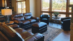 A spacious living room with a leather sofa, a coffee table, and a rug. Large windows provide a view of the outdoors. - 4 bedrooms 3 bath close to downtown and RMNP- beautiful! (Estes Park)