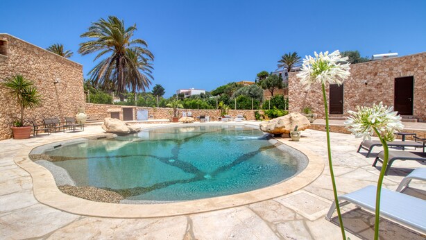A swimming pool with stone walls and palm trees, surrounded by lounge chairs and a stone building. - Il Corallo - Bilo 2 IV (Lampedusa)
