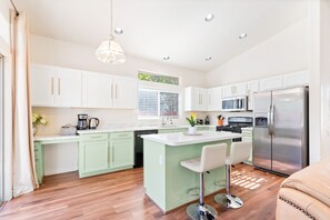 A modern kitchen with white cabinets, a stainless steel refrigerator, a large island with bar stools, and a window with blinds.