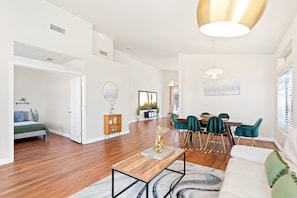 A spacious living room with a wooden floor, a white sofa, a dining area with teal chairs, and a large window.