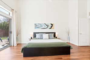 A modern bedroom with a large bed, a view through a sliding glass door, and a piece of abstract art on the wall.
