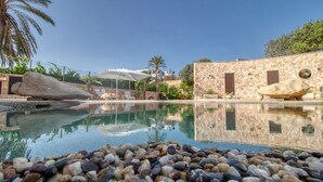 Una piscina d'aigua cristal·lina envoltada de petits pedres, amb gandules i ombrelles, i un edifici de pedra amb una porta i una finestra.