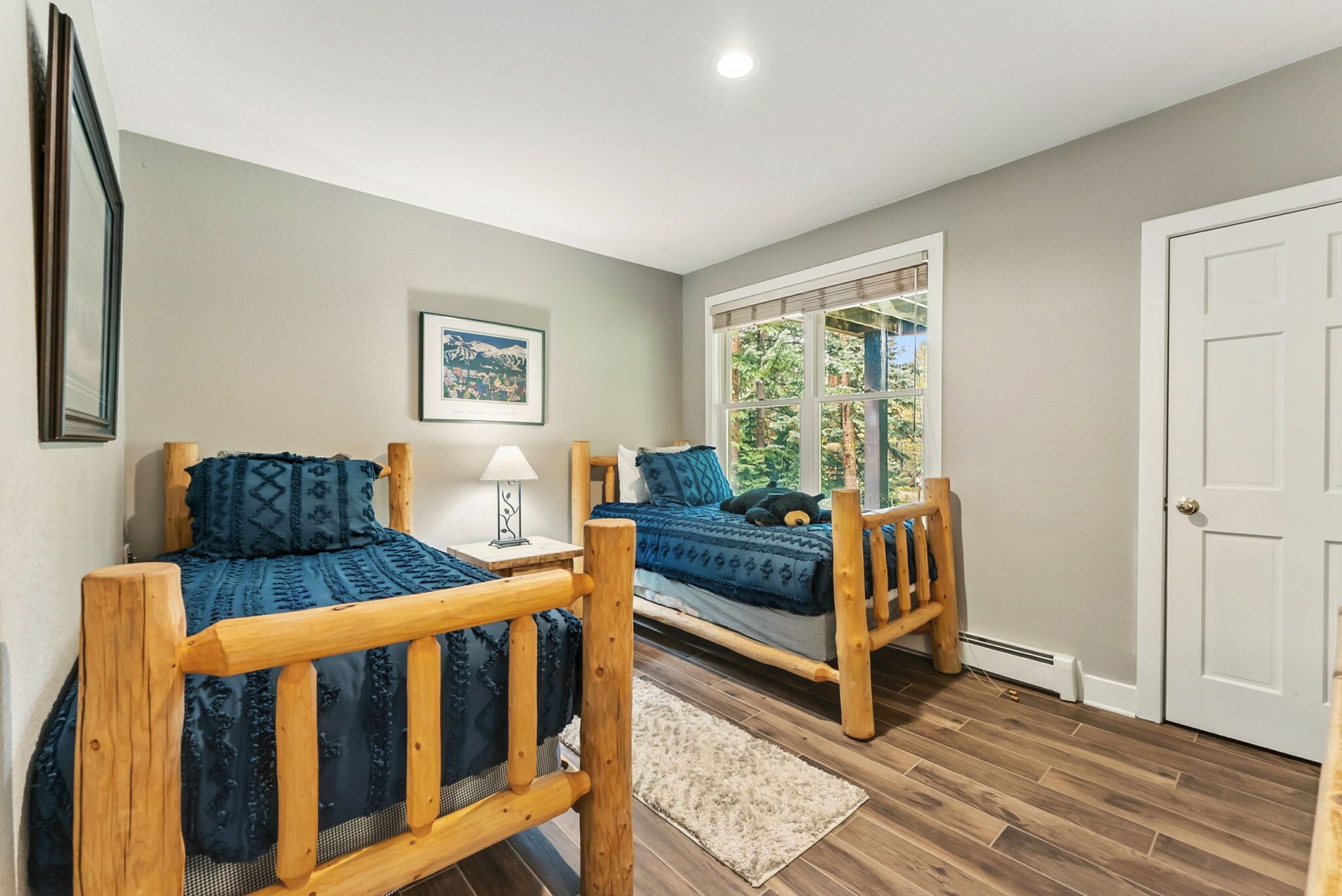 A bedroom with two wooden beds, a window with a view of greenery, a white door, and a framed picture on the wall.