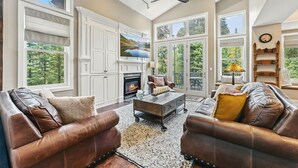 A spacious living room with leather sofas, a fireplace, large windows, and a ceiling fan.