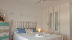 A neatly made bed with white linens and blue pillows, a wooden dresser with drawers, a nightstand with a lamp, and a large wardrobe with glass doors. - EL CABO III        . (San José)