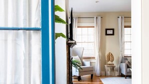 Una habitación con piso de madera, una puerta azul, un sillón gris, una mesa pequeña con un objeto decorativo y una ventana con cortinas.