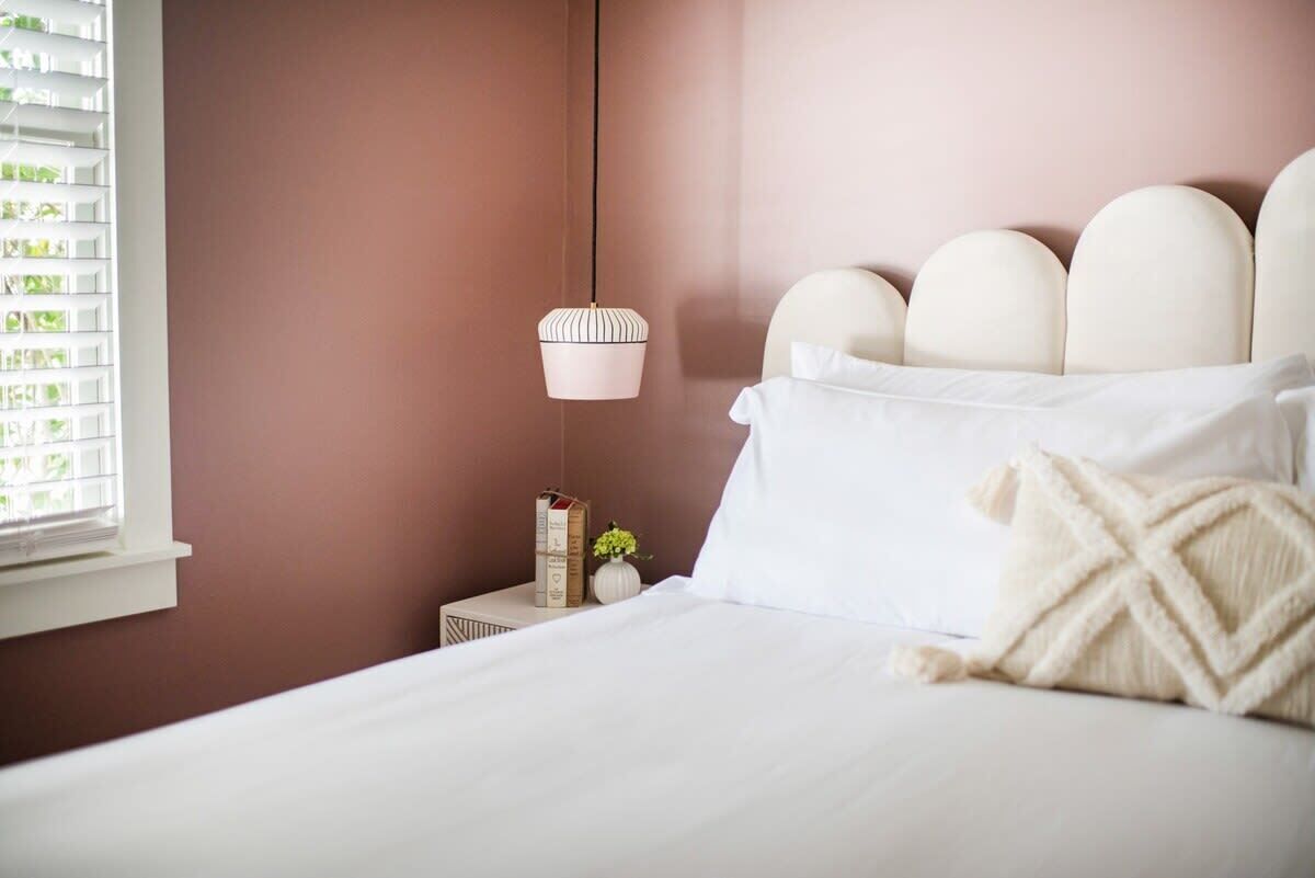 A neatly made bed with white linens and a decorative pillow, a bedside table with a lamp, and a window with blinds.