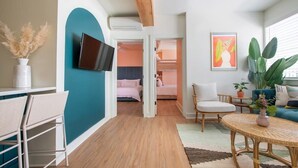 Une chambre d’hôtel moderne avec un mur d’accent de couleur turquoise, un téléviseur à écran plat, un lit avec une literie rose, un coin salon avec une petite table ronde en osier et une grande plante.