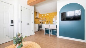 Une cuisine moderne avec des carreaux jaunes, un four encastré et un coin repas blanc agrémenté d’un téléviseur fixé sur un mur de couleur turquoise.