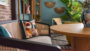 A cozy seating area with a round wooden table, a woven sofa, and a decorative wall with plants and wall art. - Soho House | Prime Location, Walk to Everything! (Charleston)