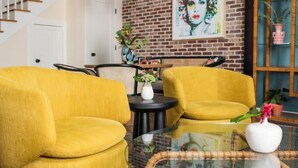 A room with a brick wall, a staircase, yellow armchairs, a round glass table, and a framed artwork. - Soho House | Prime Location, Walk to Everything! (Charleston)