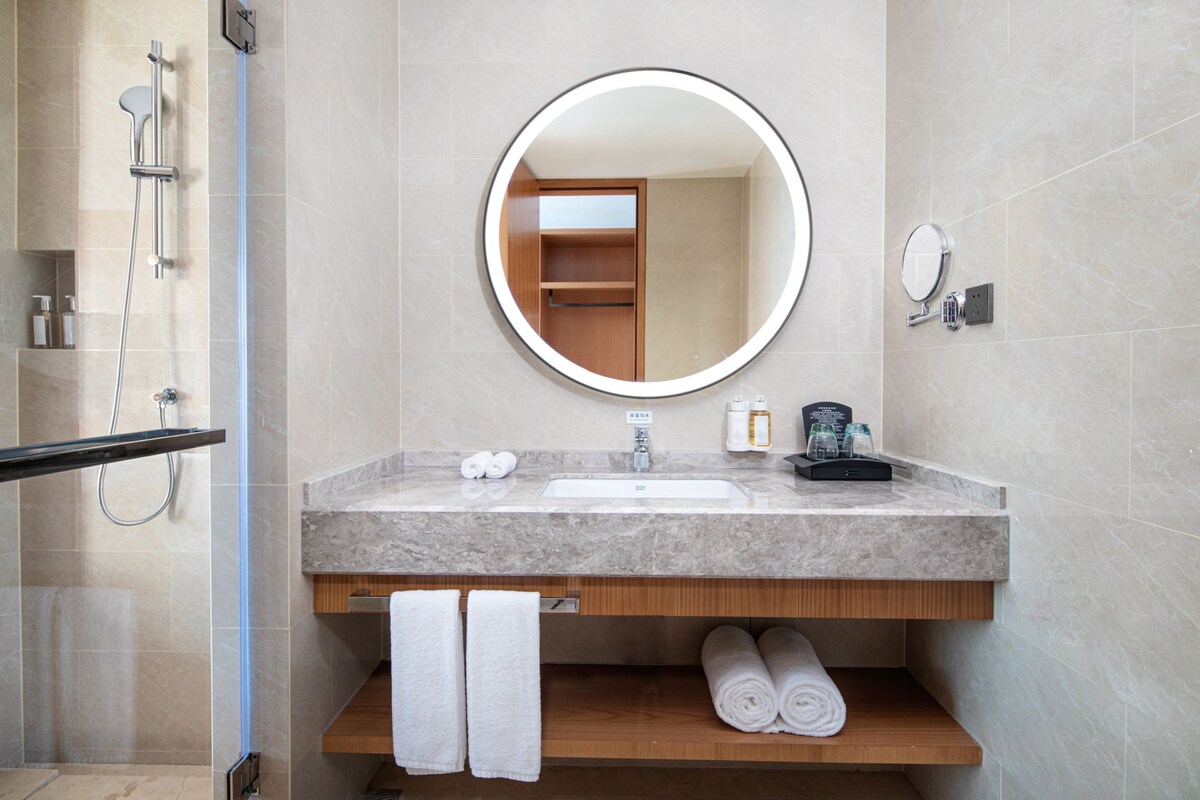 A modern bathroom with a marble sink, a round mirror, a shower, and neatly folded towels.