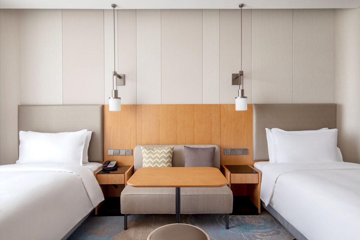 A hotel room with two beds, a wooden headboard, bedside tables, a small table, and pendant lights.