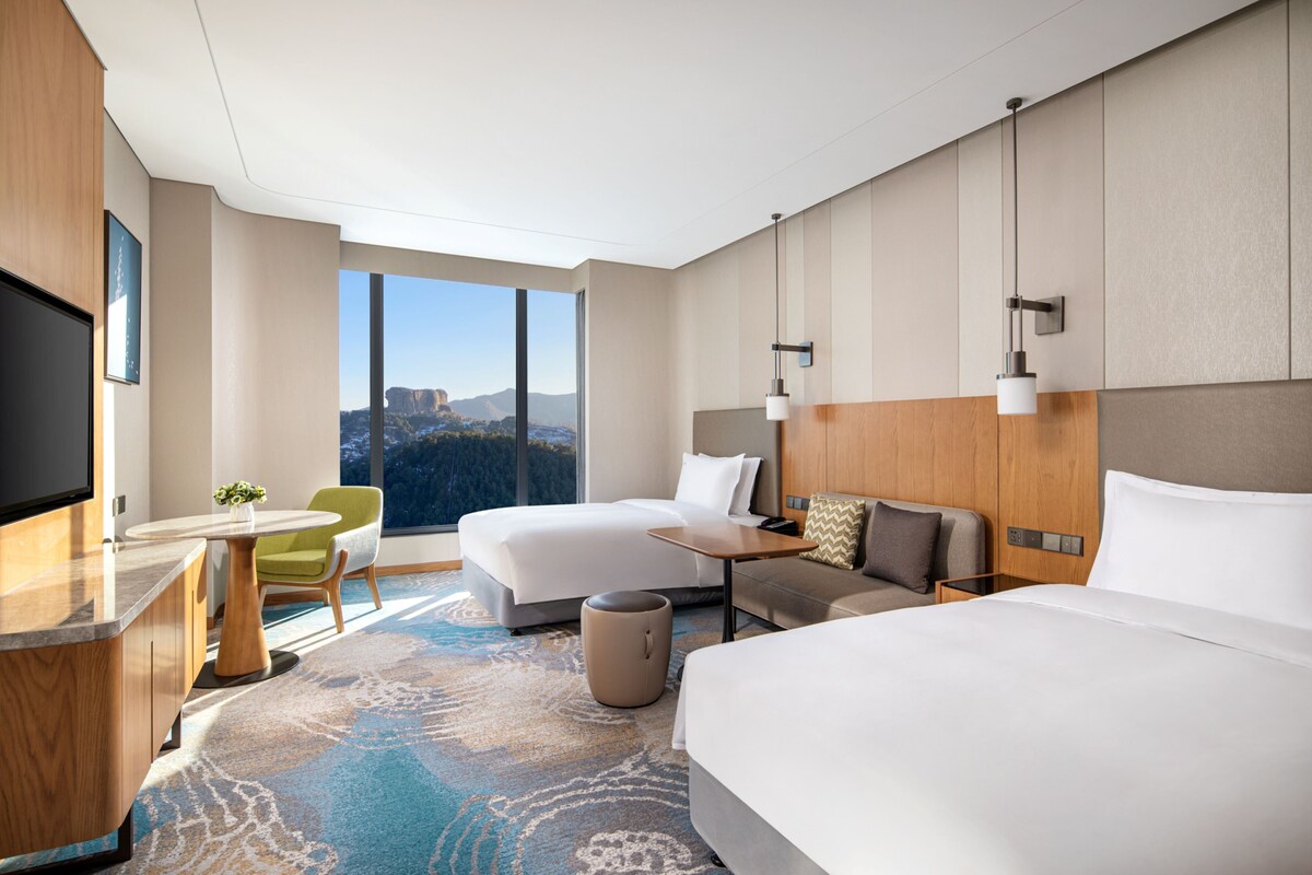 A hotel room with two beds, a desk, a chair, a TV, and a large window offering a mountain view.