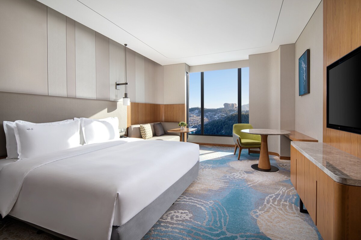 A modern hotel room with a large bed, a seating area with a table, a TV, and a view of a mountain.