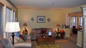 A living room with a sofa, coffee table, and lamps. There are two picture frames on the wall and a window with blinds. - Spacious House near Lake and Snowmobile Trails (Piseco)