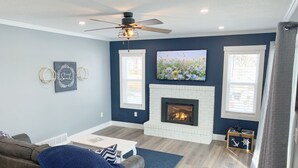 A living room with a fireplace, a ceiling fan, and a television mounted on the wall. - Beachfront Cabin with Lake Views & Harry Potter Room (Annandale)