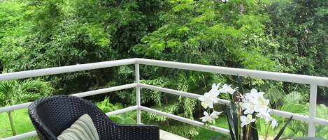 A balcony with a wicker chair and a glass table, surrounded by lush greenery.