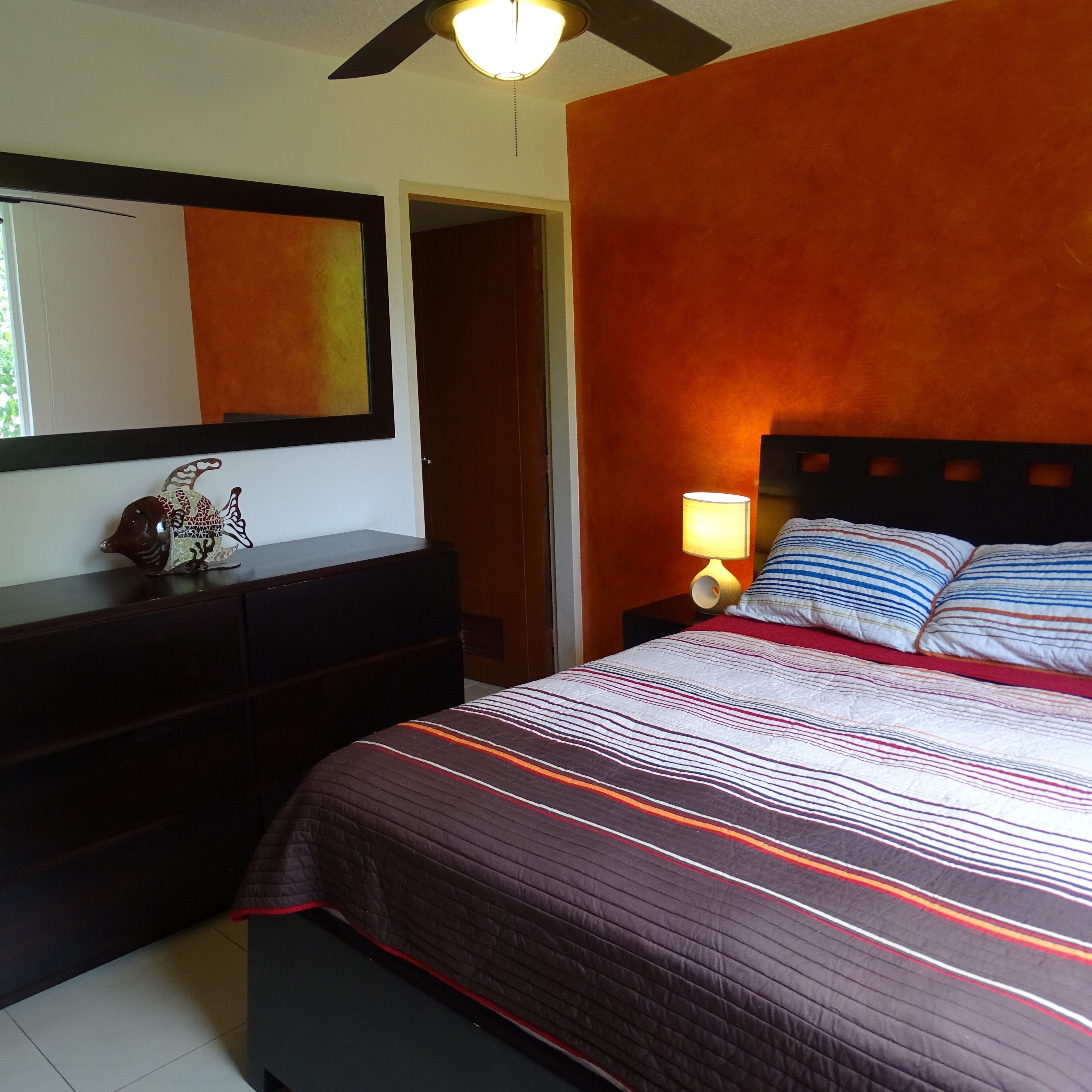 A bedroom with a bed, a dresser, a nightstand, a lamp, a ceiling fan, and a door.