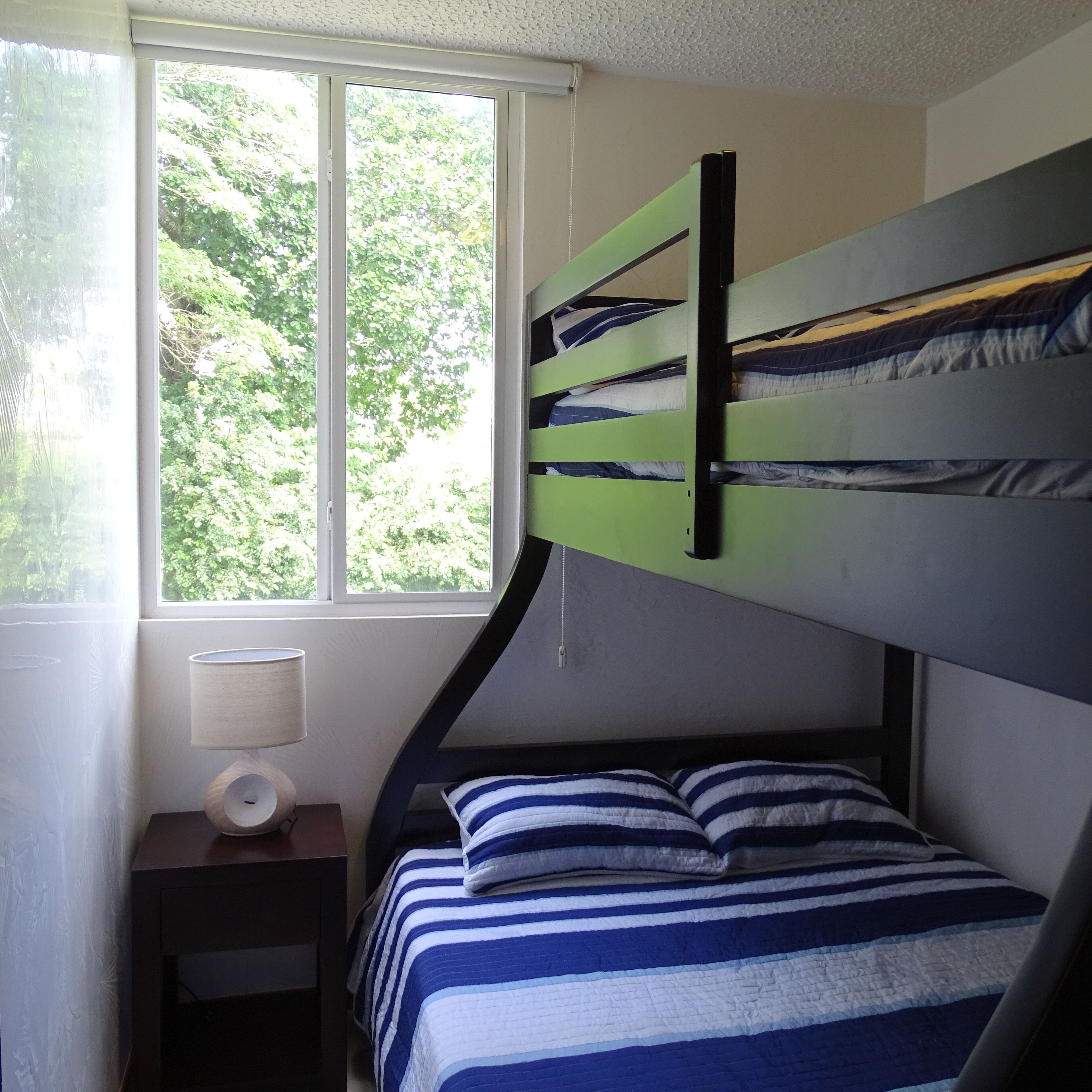 A bedroom with a bunk bed, a bedside table with a lamp, and a window with a view of greenery.