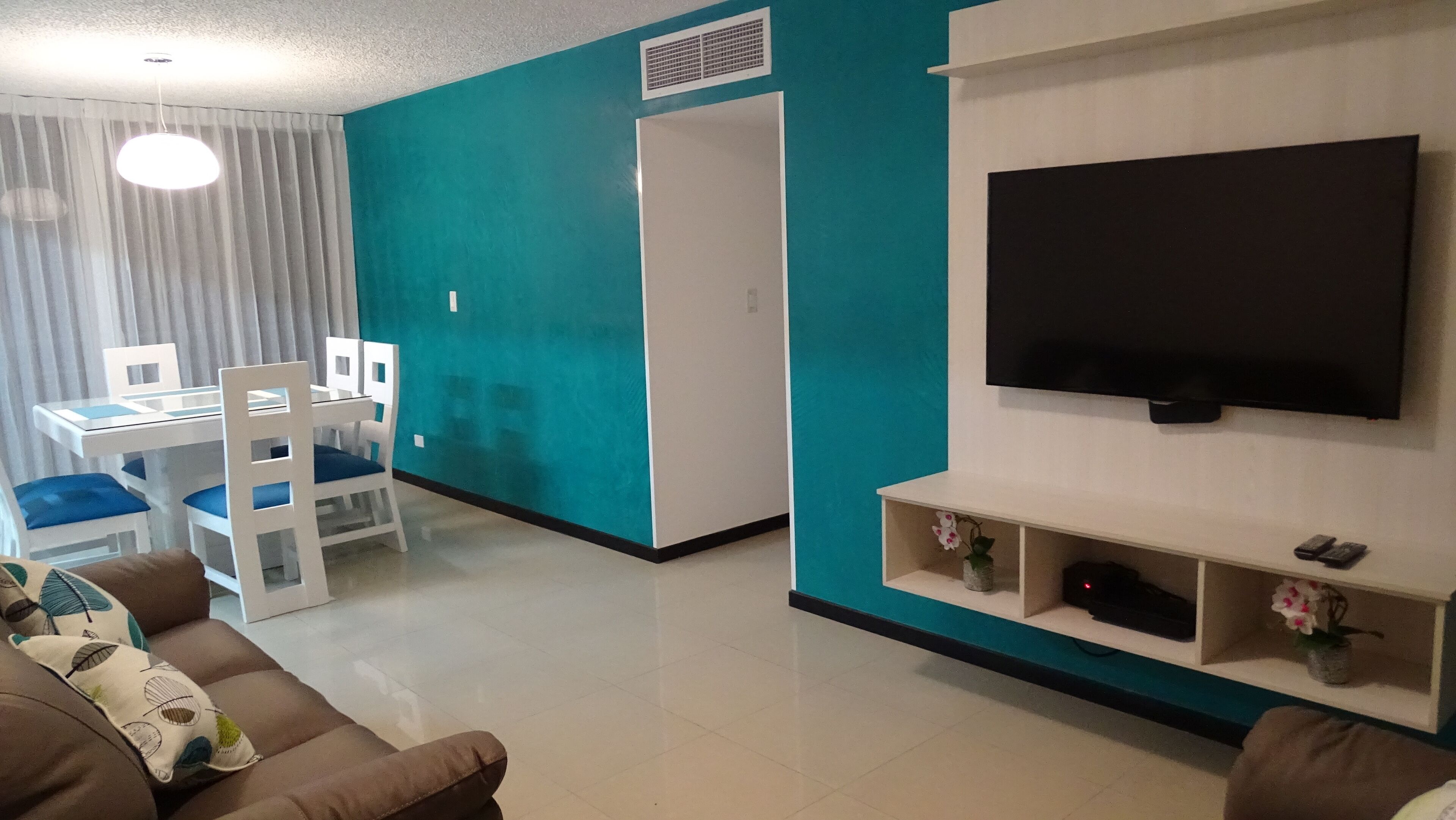 Un salon avec un mur d’accent de couleur turquoise, un téléviseur à écran plat fixé au mur, un coin repas avec une table et des chaises blanches, et un canapé marron agrémenté de coussins à motifs.