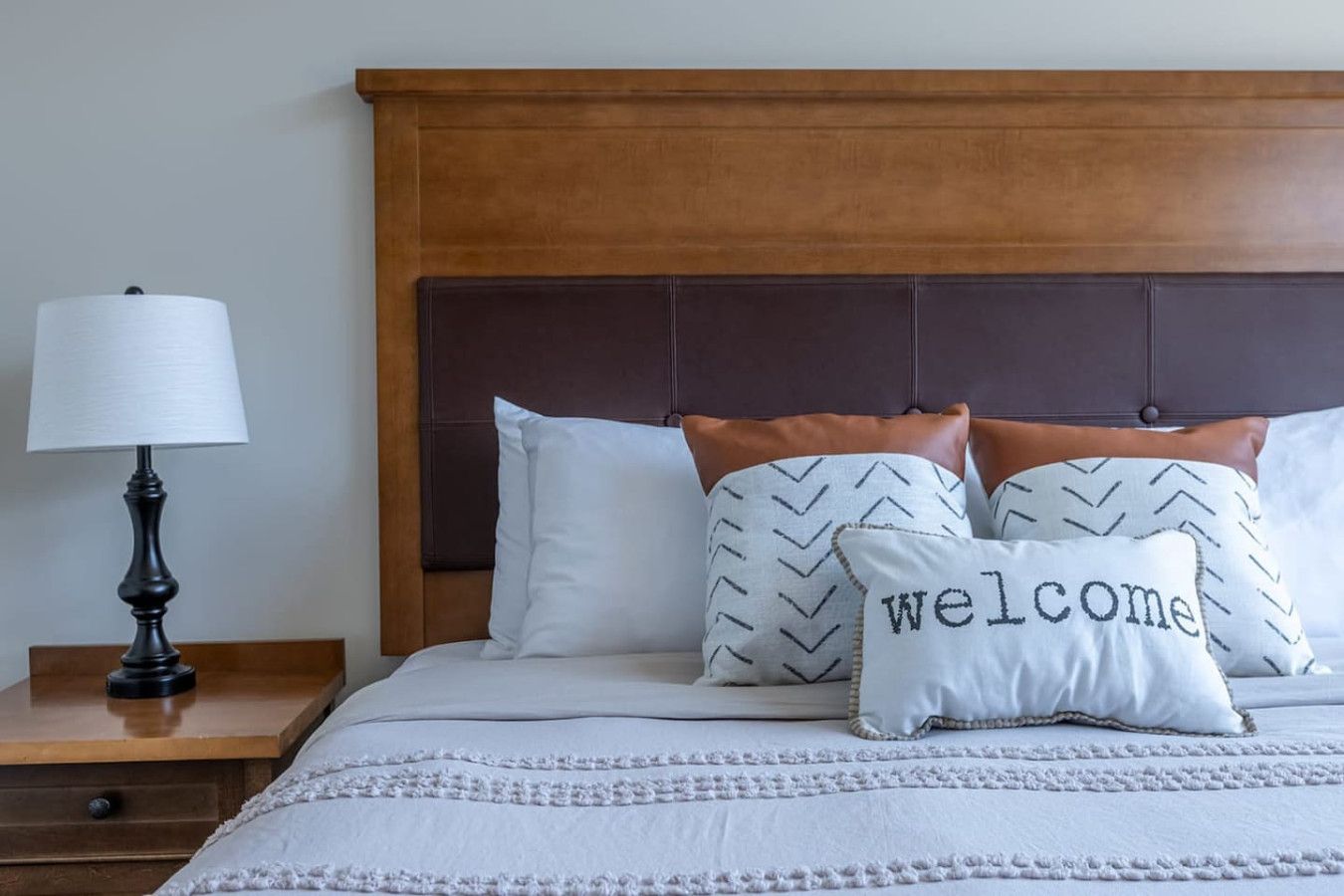 Cama bem arrumada com cabeceira, luminária de cabeceira e almofadas decorativas, incluindo uma com a inscrição 'welcome'.