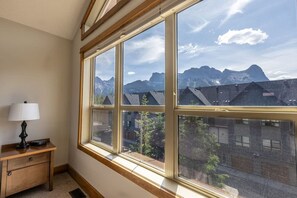 Una habitación con ventanas amplias que ofrecen vistas a la montaña, una mesita de noche de madera con lámpara y suelo alfombrado.