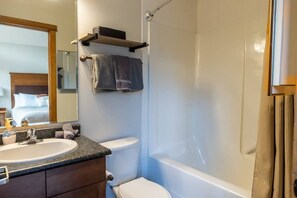 Un baño con lavabo, inodoro y bañera. Hay un perchero sobre el inodoro y un estante con toallas.