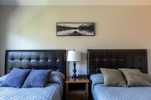 Una habitación de hotel con dos camas, cada una con cabecero y almohadas, una mesita de noche con lámpara y un cuadro en la pared.