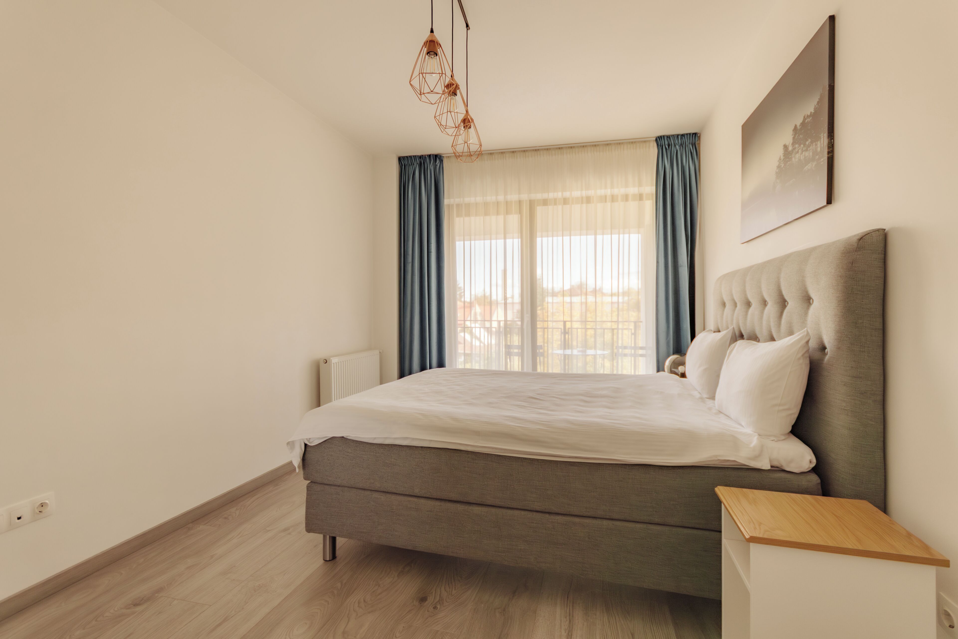A bedroom with a bed, a bedside table, a hanging light fixture, a window with curtains, and a wall-mounted picture.