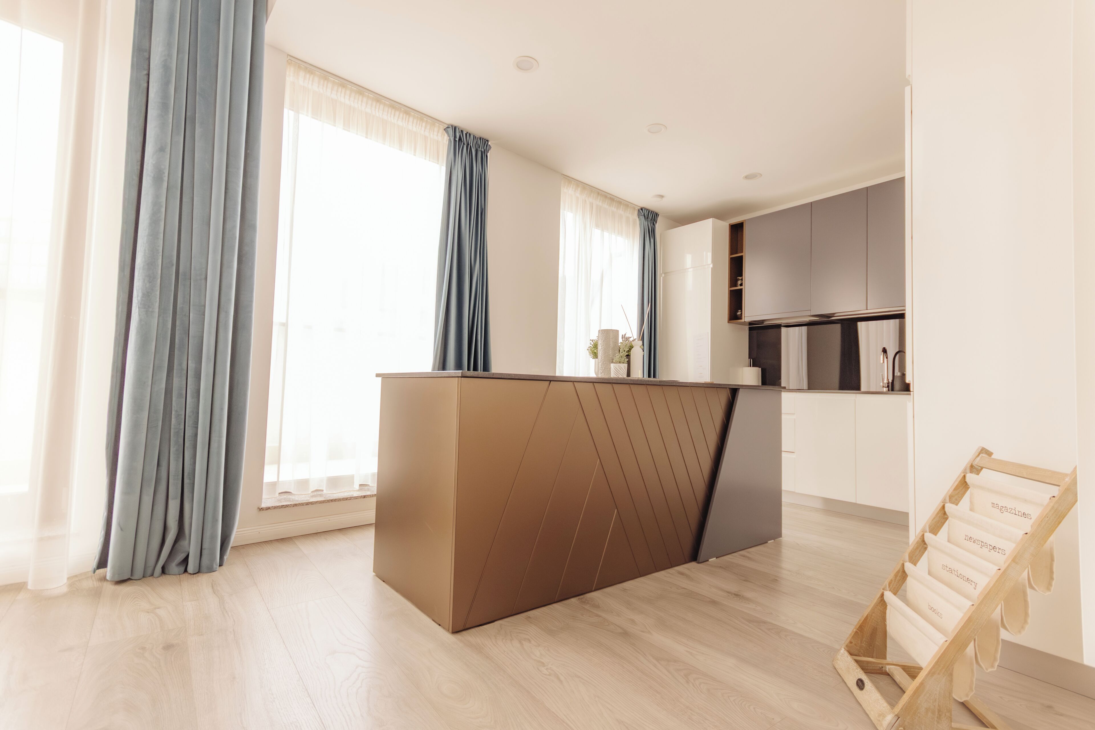 A modern kitchen with a central island, light wooden flooring, and large windows with curtains.
