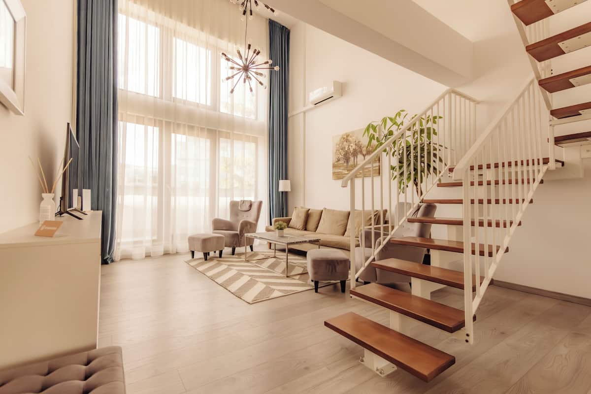 Una sala de estar moderna con una escalera, un sofá, una mesa de centro y un amplio ventanal con cortinas.