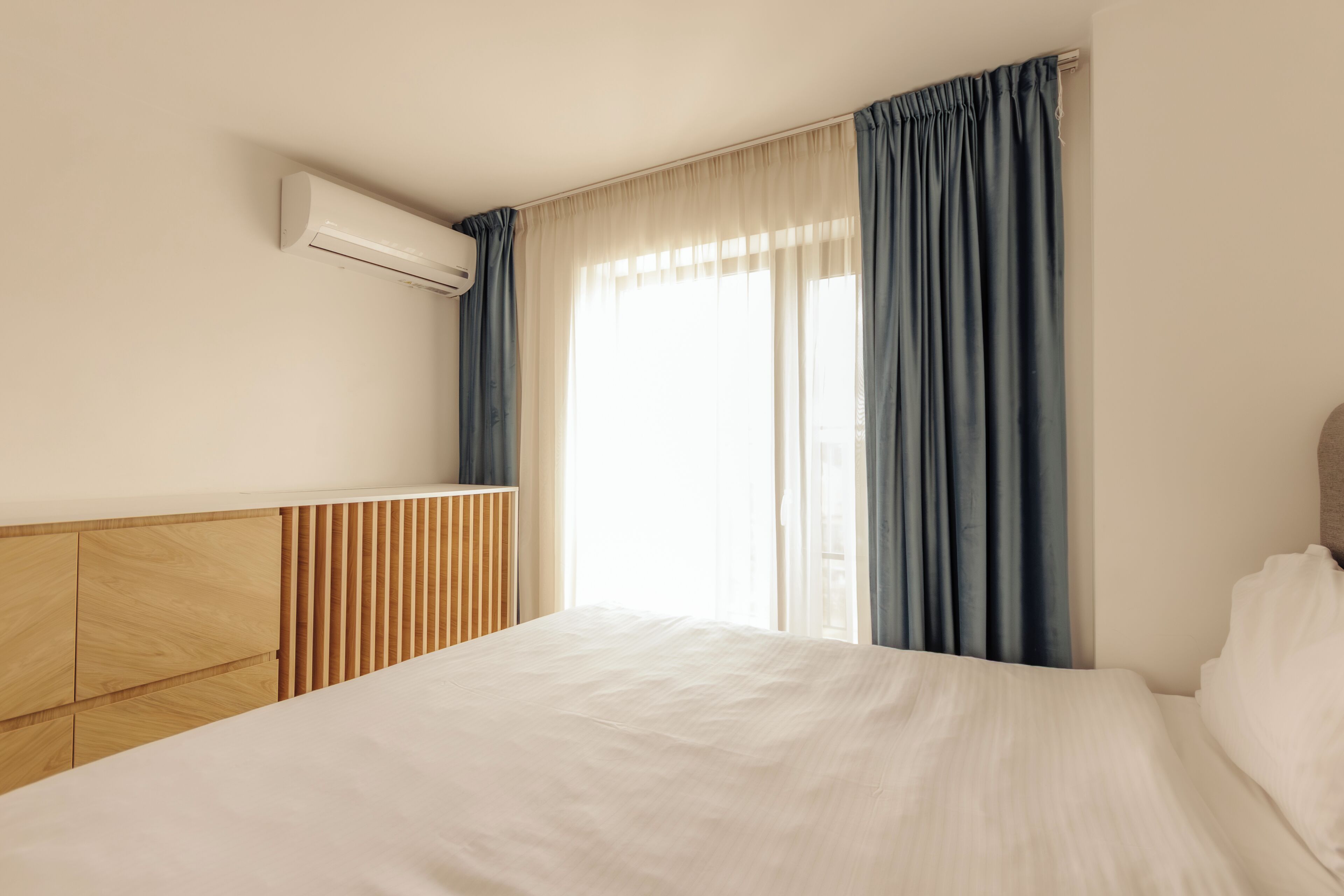 A hotel room with a bed, a wooden headboard, a built-in air conditioning unit, and a window with curtains.