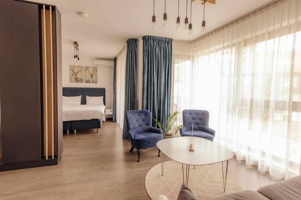 Una habitación de hotel moderna con una cama, dos sillones, una mesa redonda y un gran ventanal con cortinas.