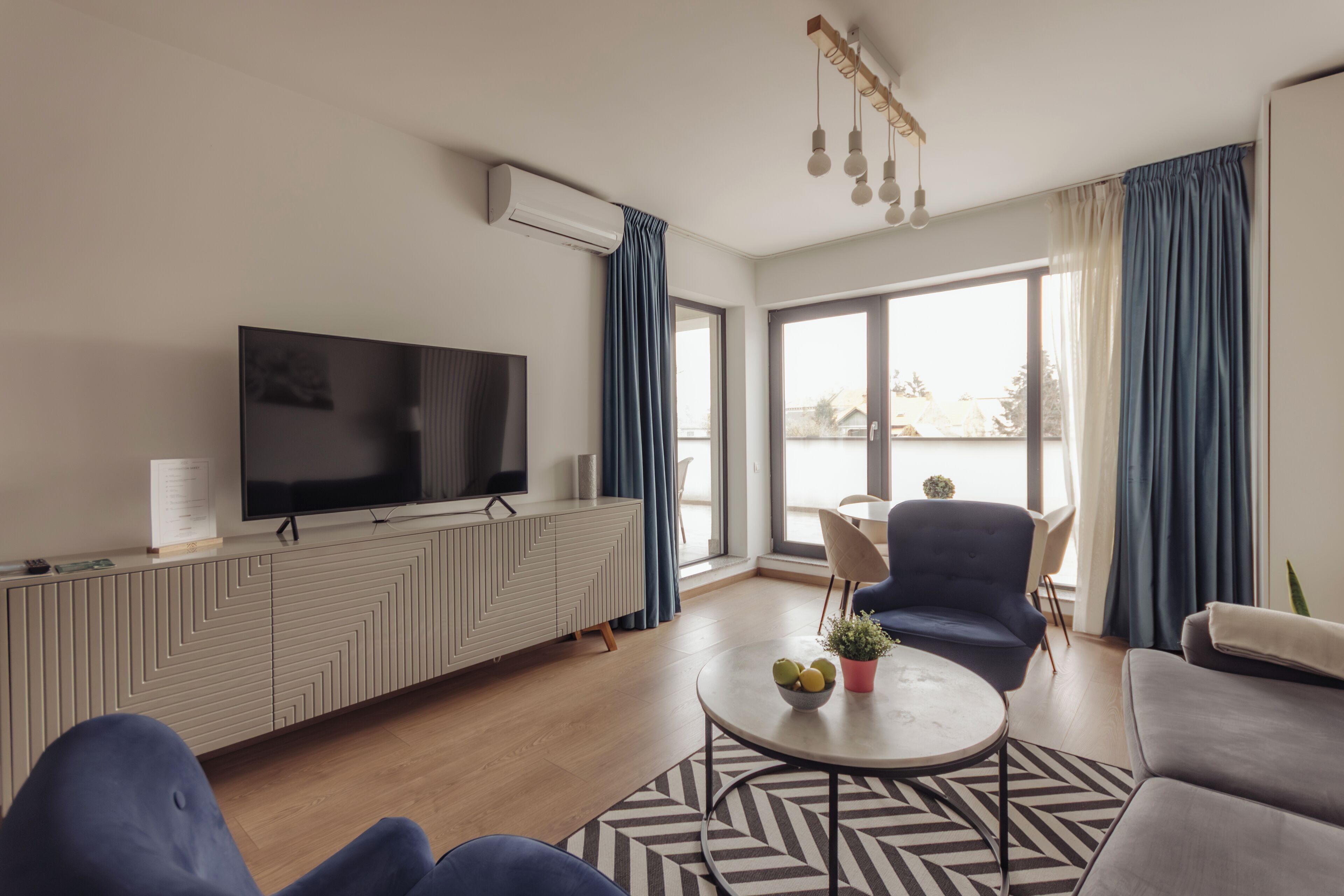 A modern living room with a flat-screen TV, a sofa, a coffee table with fruit, and a view of the outdoors.