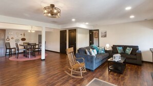A spacious living room with a wooden floor, a blue sofa with cushions, a rocking chair, and a dining area with a table and chairs. - Lakefront duplex property, nicely finished. 2 bedroom and queen size hide-a-bed. (Divide)