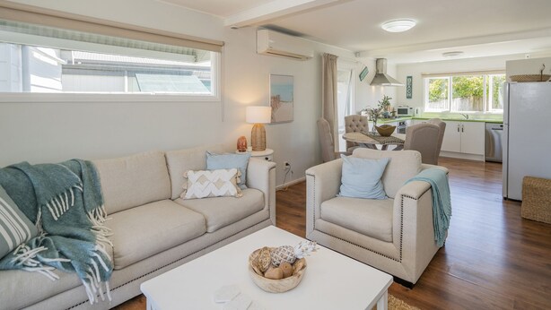 Una sala de estar con sofá, sillón y mesa de centro. Se ve una cocina al fondo, con una mesa de comedor y sillas.